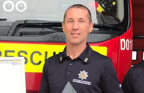 Mark Harrison, an emergency response instructor, stands in front of a red fire and rescue vehicle wearing a dark uniform adorned with a badge and text. He proudly holds a framed certificate or award, symbolizing his dedication to the fire and rescue services.