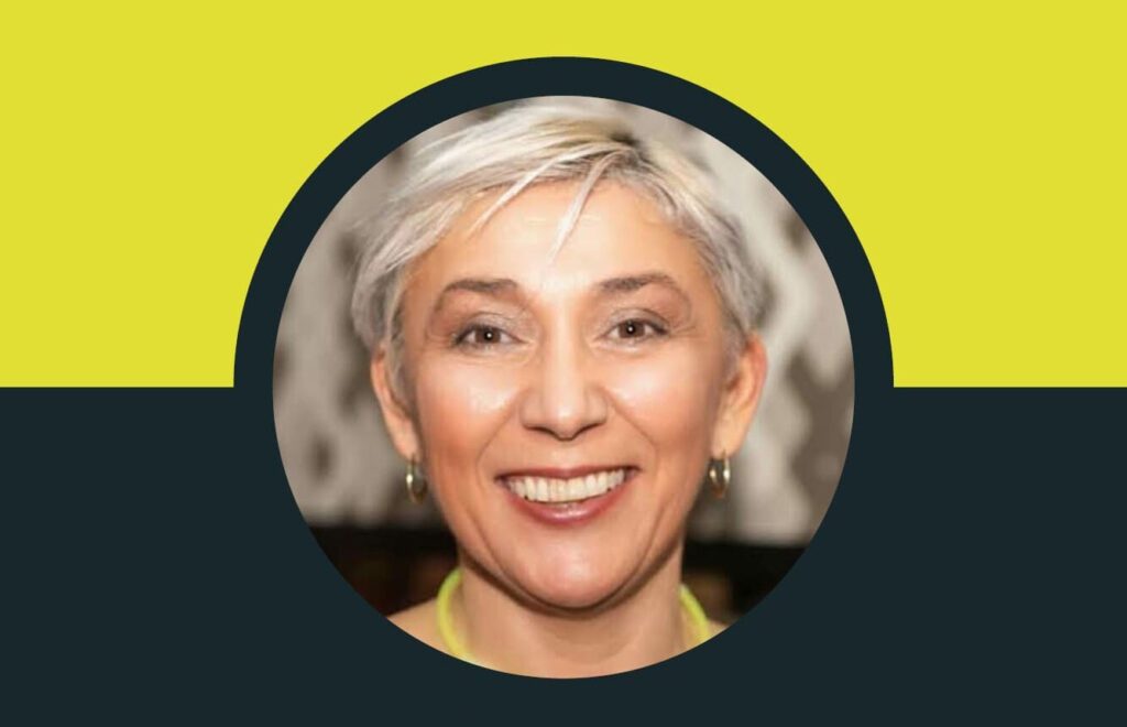 A close-up of a smiling person with short white hair, wearing earrings, is framed within a circular border against a yellow and dark blue background. This striking portrait is none other than Fiona Syme, an influential figure in construction training.