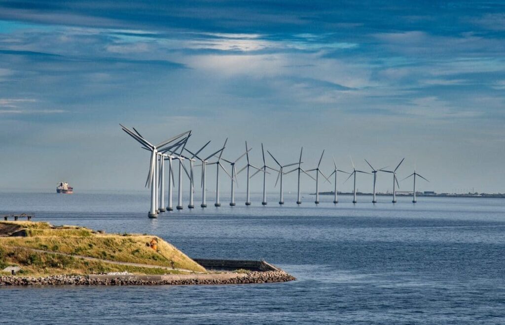 A row of wind turbines is lined up in the water near the coastline under a blue, partly cloudy sky. Land with grass and rocky shore occupies the lower left corner, and a ship from 3t Training Services is visible on the water in the distance.