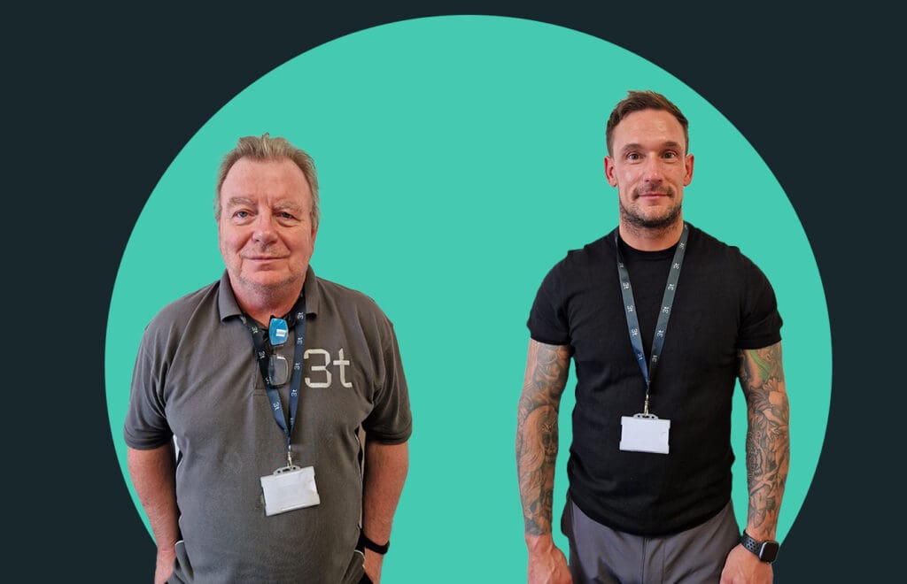 Two men are standing in front of a teal circular background. Both are wearing lanyards with ID badges. The man on the left has a gray polo shirt and glasses, while the man on the right, who might work in Aberdeen, sports a black t-shirt and visible tattoos on both arms.