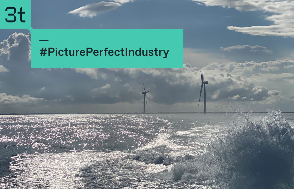 Ocean waves ripple under a cloudy sky with sunlight reflecting on the water. Two wind turbines stand on the horizon, tirelessly spinning. A teal-colored caption in the top left corner reads "#PicturePerfectIndustryTraining.