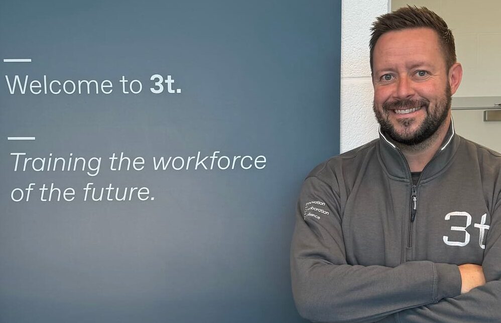 A man wearing a grey jacket with "3t" on it stands next to a sign that reads, "Welcome to 3t. Training the workforce of the future." He is smiling and has his arms crossed, embodying confidence as the new centre manager of 3t in Teesside.